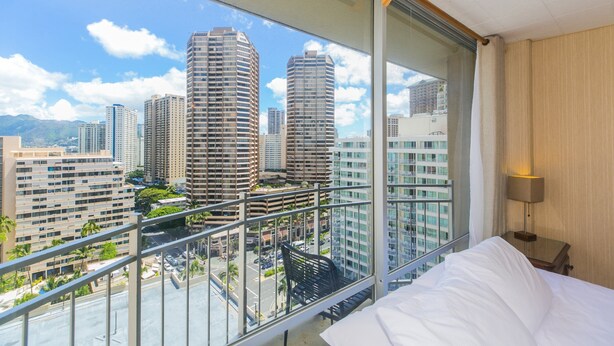 A hotel room with a balcony overlooking a cityscape with tall buildings and a clear sky.
