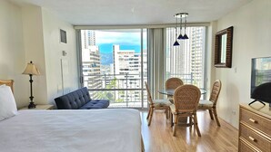 A hotel room with a large bed, a dining table with chairs, a television, and a balcony with a view of the city.
