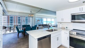 A modern kitchen with a dishwasher, stove, and microwave. There is a dining area with blue chairs and a table. The room has large windows with a view of buildings and the ocean. - Desirable "44"; Stack At The Ilikai Hotel.  Unobstructed Ocean Views! (Honolulu)
