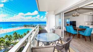 A balcony with a glass table and chairs, a dining area with blue chairs, and a view of a beach and ocean. - Desirable "44"; Stack At The Ilikai Hotel.  Unobstructed Ocean Views! (Honolulu)