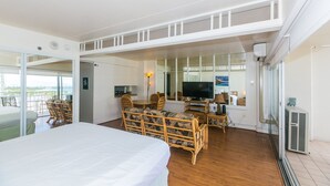 Habitación de hotel con cama, zona de comedor con muebles de mimbre, televisor y un amplio ventanal con vistas.