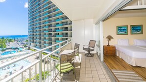 Een balkon met uitzicht op een hotelzwembad en de oceaan, een tafeltje met stoelen en een slaapkamer met een bed en nachtkastje.
