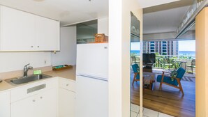 Een moderne keuken met witte kasten, een roestvrijstalen spoelbak en een kleine koelkast. Er is een balkon met een houten vloer, blauwe stoelen en een tafel met een televisie.