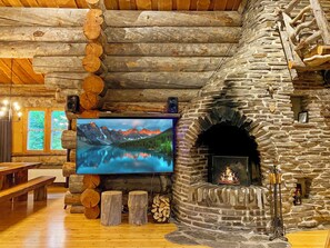 L’intérieur d’un chalet accueillant, avec une cheminée en pierre, des rondins de bois, un téléviseur et une vue sur un paysage montagneux.
