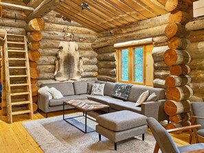 A log cabin interior with a sectional sofa, a wooden coffee table, a ladder, and a window with a view of trees.