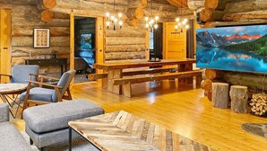 A log cabin interior with a wooden dining table, benches, and a large flat-screen TV displaying a mountain lake.