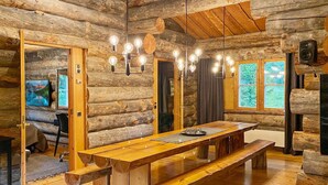 A log cabin interior with a wooden dining table, benches, a chandelier, and a television on the wall.