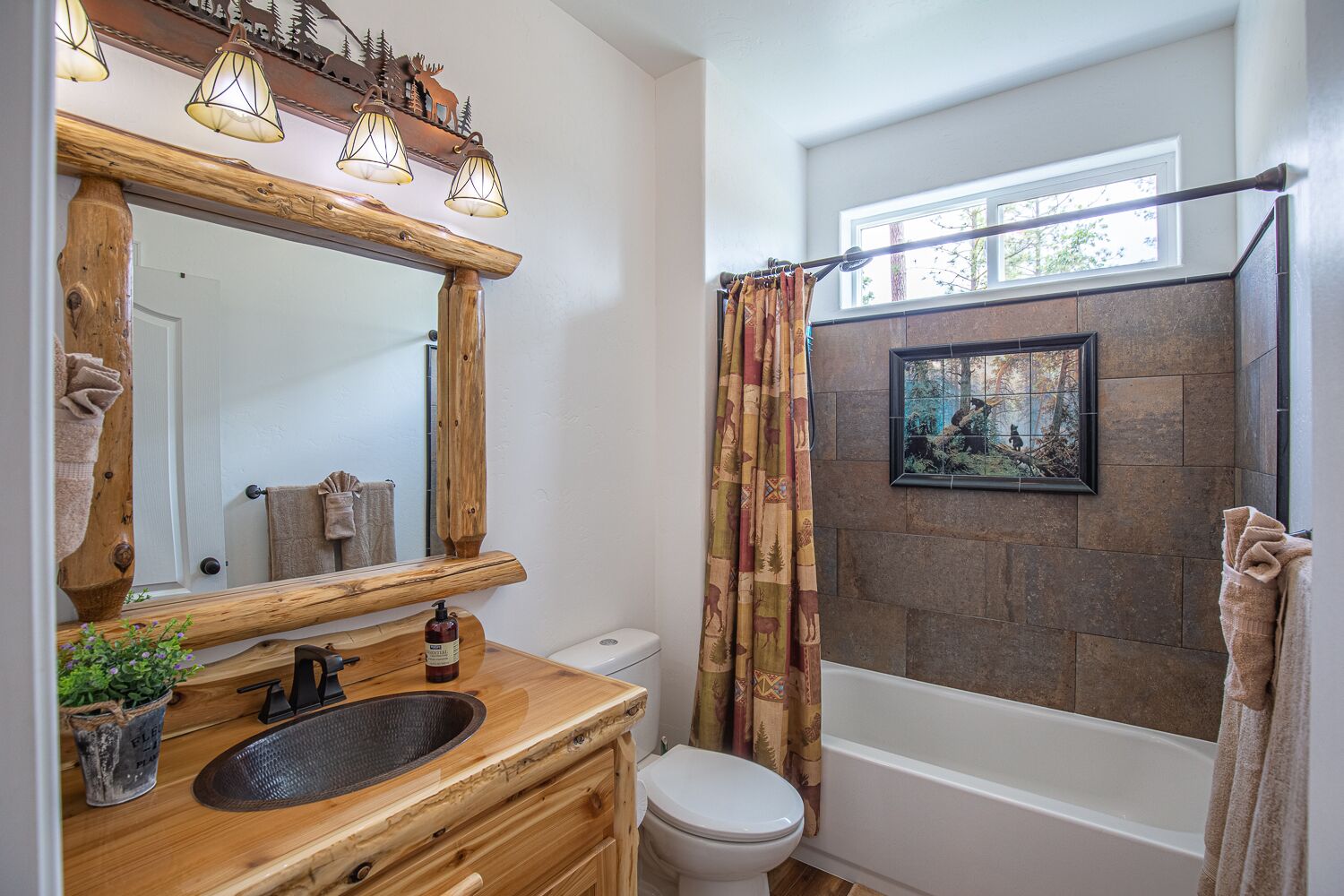 Une salle de bain avec un meuble-lavabo en bois, un grand miroir, des toilettes, une baignoire et un rideau de douche orné d’un motif représentant une forêt.