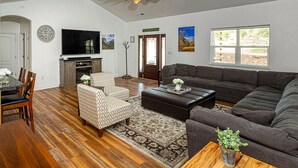 Uma sala de estar espaçosa com um sofá cinza escuro, poltronas estampadas, uma mesa de jantar de madeira com cadeiras, uma televisão de tela plana e uma grande janela com vista para áreas verdes.
