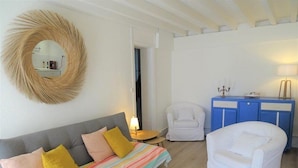 A room with a grey sofa, a colorful striped bedspread, a white armchair, a blue cabinet, and a round mirror on the wall.