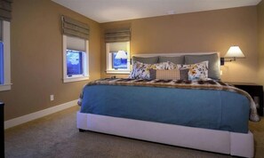 A bedroom with a large bed, two windows with blinds, a bedside lamp, and a carpeted floor.