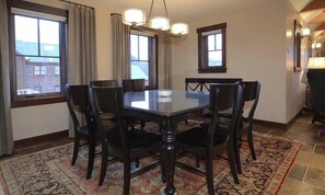 Een eetkamer met een ronde tafel, zes stoelen, een kroonluchter en uitzicht op een gebouw door grote ramen.