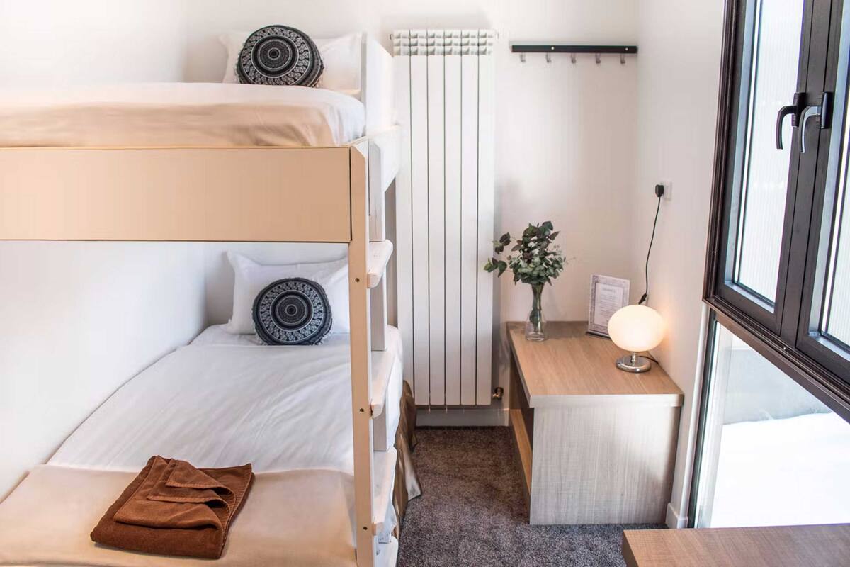 A compact hotel room with a bunk bed, a small desk with a lamp, and a radiator.