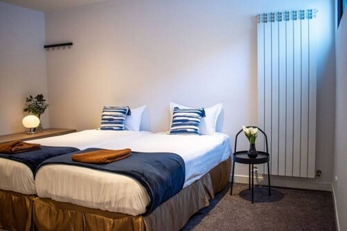 A hotel room with a bed, a nightstand with a lamp, a chair with a vase, and a radiator.