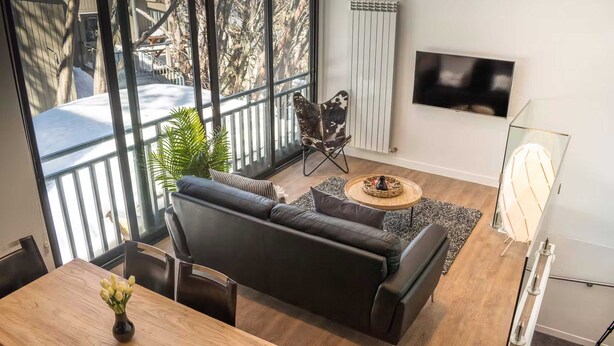 Una sala de estar moderna con un sofá de cuero negro, una mesa de comedor de madera con un jarrón de flores, una mesa de centro de vidrio, un televisor montado en la pared y un radiador.