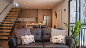 Una sala de estar moderna con un sofá de cuero negro, un comedor con cocina al fondo y una escalera que sube a un nivel superior.