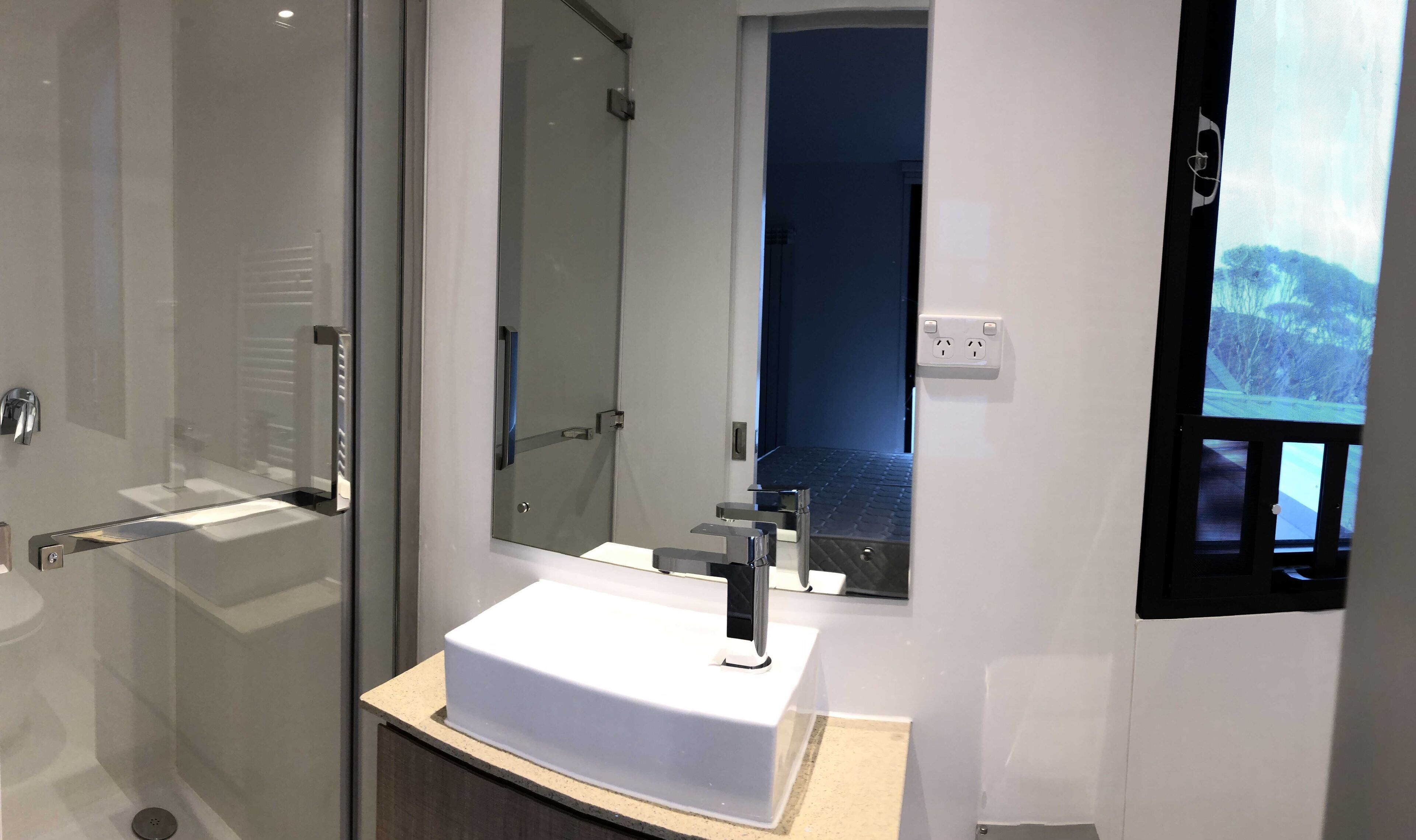 Une salle de bain moderne avec une douche à parois de verre, un lavabo blanc doté d’un seul robinet et un grand miroir.