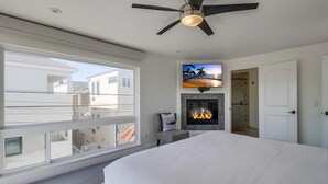 Un dormitorio con una cama grande, un ventilador de techo, un televisor sobre la chimenea y un balcón con vistas a edificios vecinos.