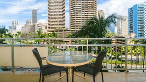 A balcony with a round glass table and two chairs, overlooking a cityscape with tall buildings and palm trees.