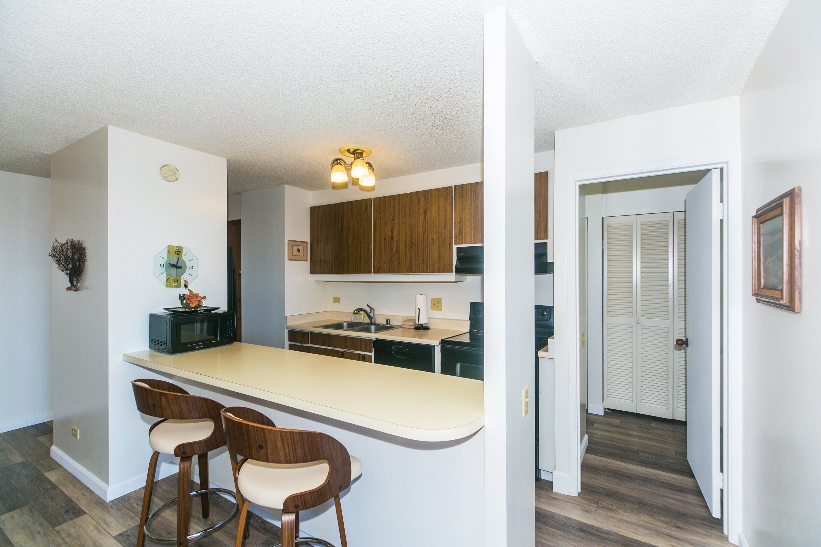 A modern kitchen with a central island, wooden cabinets, and a microwave. There are two bar stools and a small plant on the island.