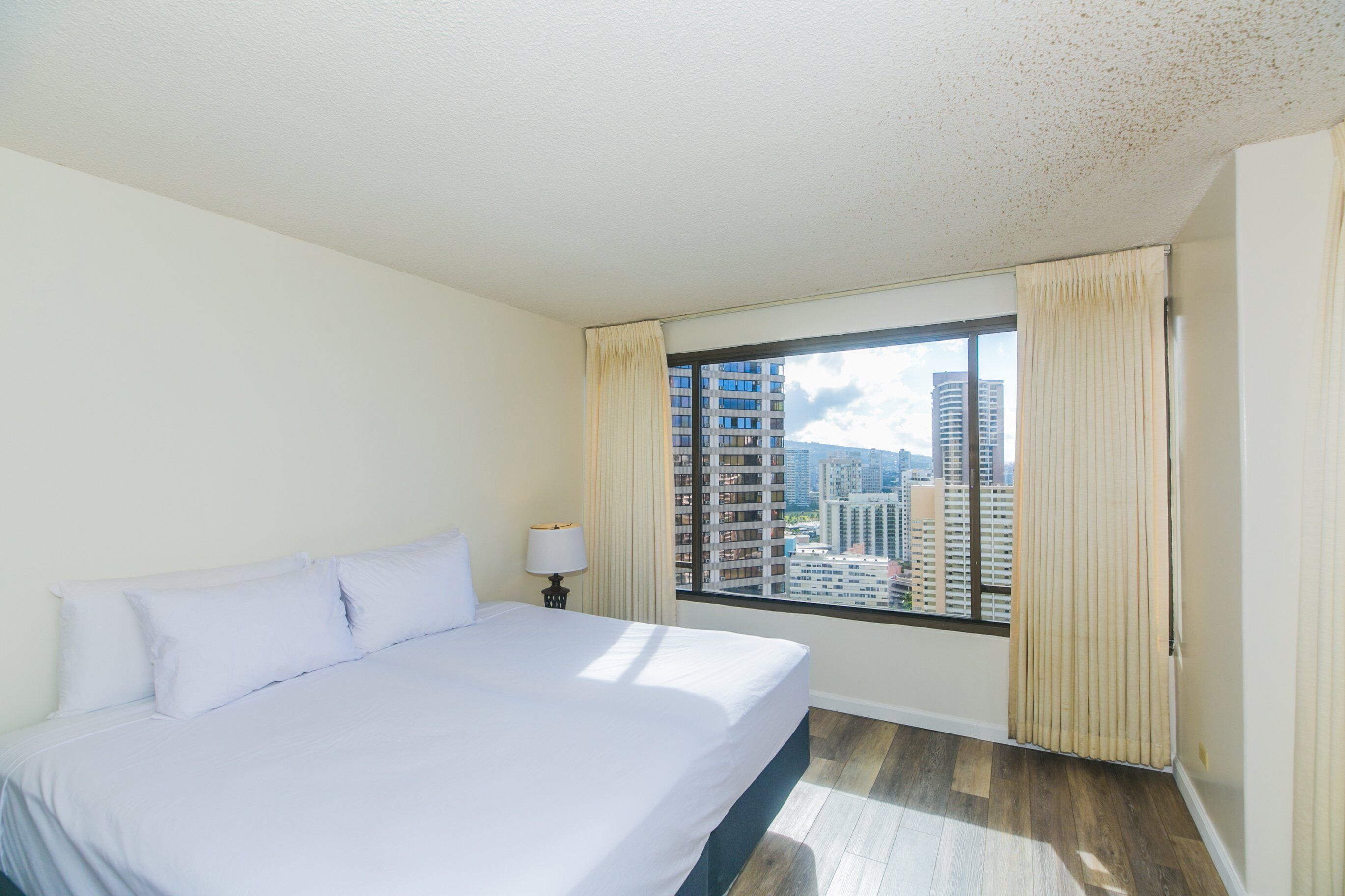A hotel room with a large bed, a window with curtains, and a view of tall buildings.