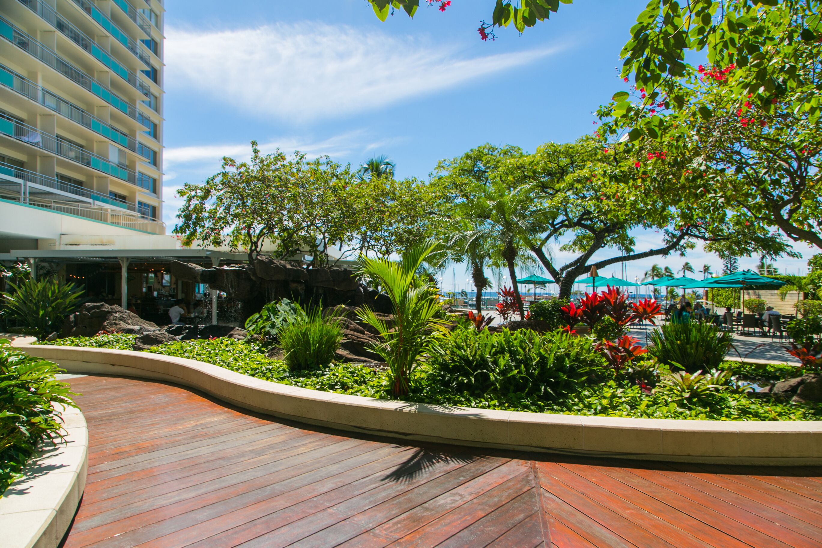 Zona esterna curata con giardino, un deck in legno curvo, piante tropicali e una zona piscina con ombrelloni.