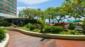 A landscaped outdoor area with a curved wooden deck, tropical plants, and a pool area with umbrellas. - Cozy 14th floor condo overlooking the surrounding Waikiki area!  Great price! (Honolulu)