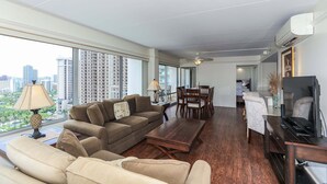 Una sala de estar moderna con sofá, mesa de centro, mesa de comedor y televisión. Amplias ventanas ofrecen vistas a la ciudad.