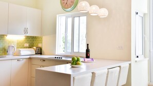 Une cuisine avec des armoires blanches, un pare-feu vert, une fenêtre donnant sur un beau paysage, une horloge et un saladier de fruits posé sur le comptoir.