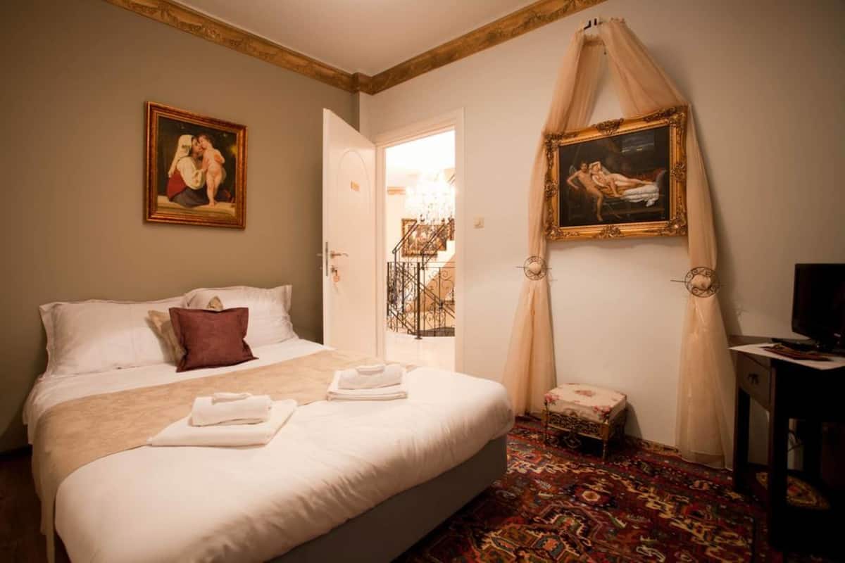 A hotel room with a large bed, a small table, a chair, and two paintings on the walls.