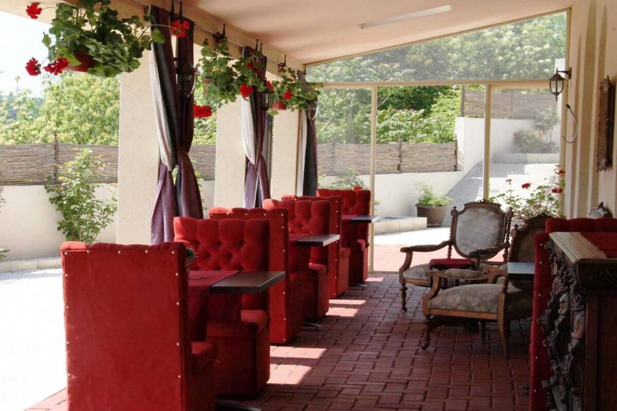 A covered outdoor seating area with red upholstered chairs and tables, a wooden chair, and hanging plants.