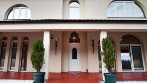 A large, two-story building with a symmetrical facade, featuring a central entrance with a decorative archway, flanked by two large windows and two potted plants.
