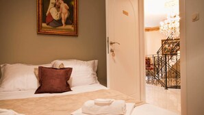 A hotel room with a bed, a painting on the wall, a chandelier, and a staircase with a wrought iron railing.