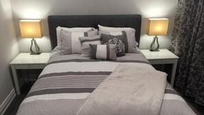 Uma cama bem arrumada com colcha listrada e várias almofadas, com duas mesas de cabeceira e abajures ao lado.