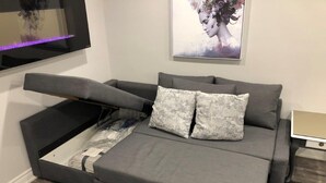 Um sof á-cama moderno com tecido cinza, duas almofadas cinzas e estrutura de madeira.