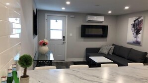 Uma sala de estar moderna com um sofá cinza, uma mesa de centro e uma TV fixada na parede. Há uma porta com janela, uma pequena planta em vaso e um vaso com flores sobre a mesa.