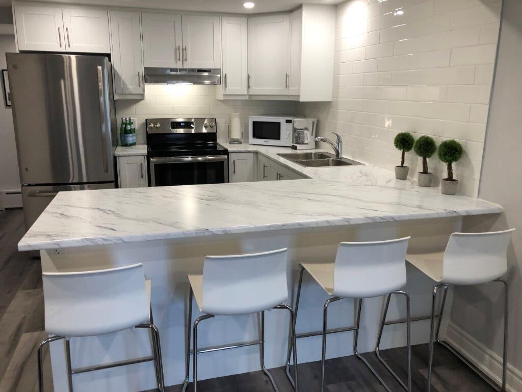 A modern kitchen with white cabinets, stainless steel appliances, a marble countertop, and four white bar stools.