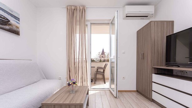 Una habitación moderna y bien iluminada con un sofá, una mesa de centro de madera con un florero, un televisor sobre un mueble bajo, y un ventanal corredero que da a un espacio exterior con una silla.