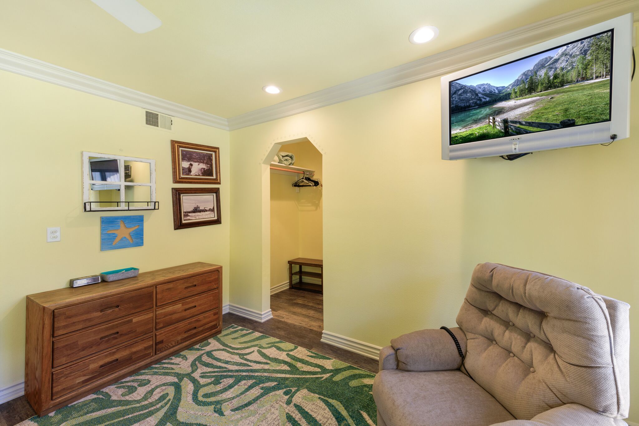 Un couloir avec une commode en bois, un fauteuil à capitons, un téléviseur fixé au mur qui diffuse un paysage pittoresque, et un tableau encadré accroché au mur.