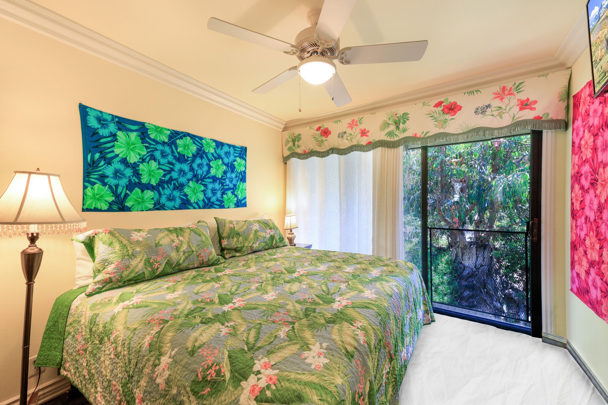 A bedroom with a bed, a ceiling fan, a large window with curtains, and a floral painting on the wall.