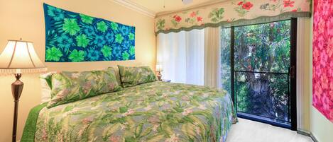 A bedroom with a bed, a ceiling fan, a large window with curtains, and a floral painting on the wall.