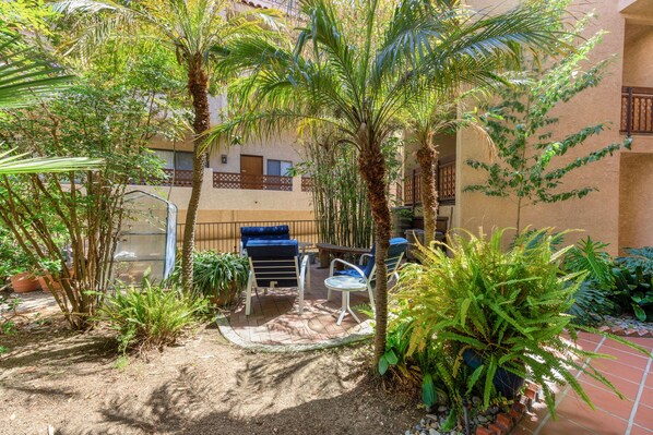 A courtyard with palm trees, a seating area with chairs and a table, and a building with balconies in the background.