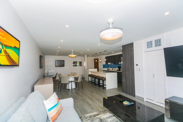 A modern living room with a sofa, a coffee table, and a dining area with chairs and a table. There is a kitchen area with a counter and stools. - Great Location In Wakiki, Walking Distance To Beach, Shopping, Dining! (Honolulu)