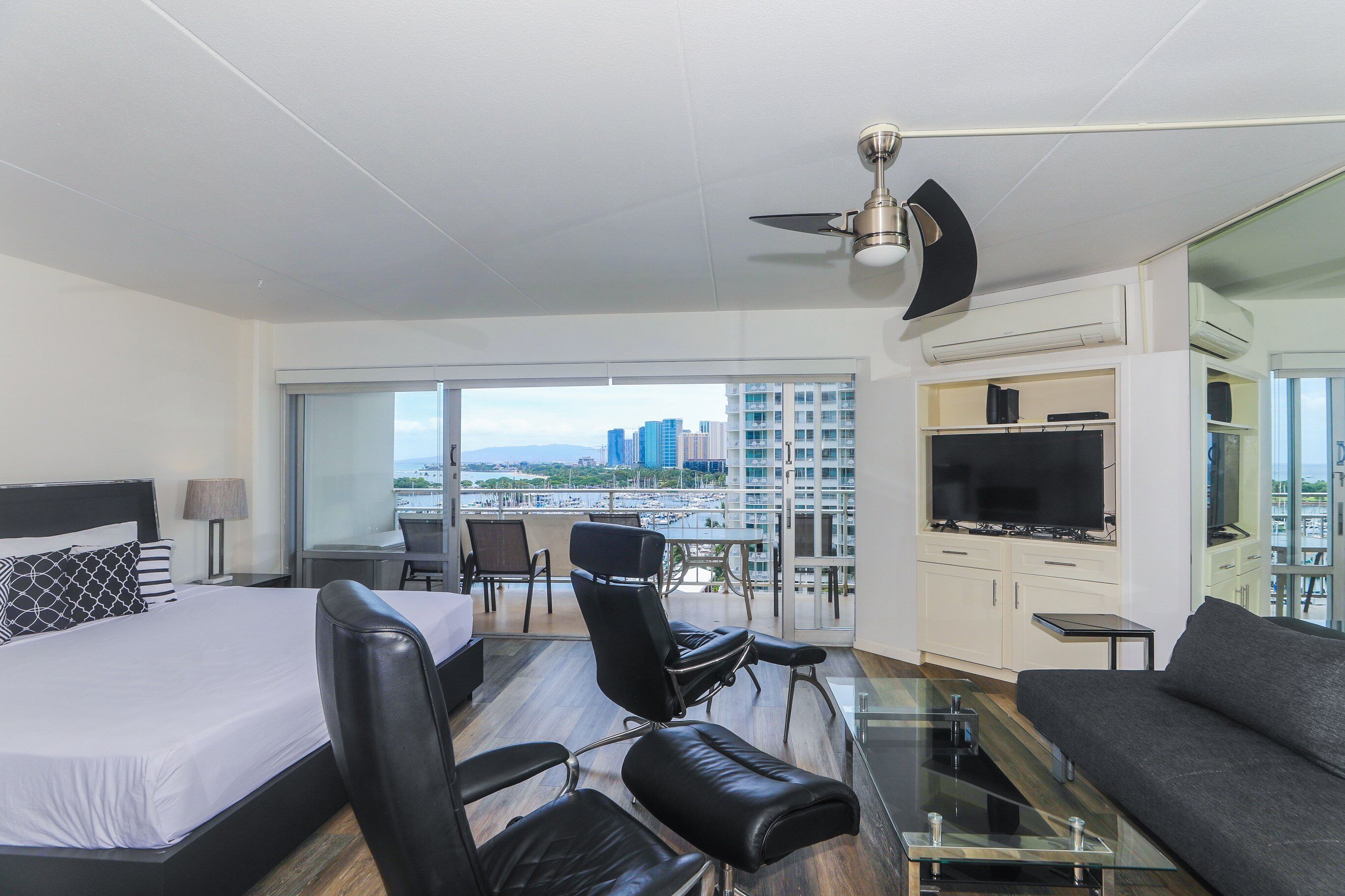 A modern hotel room with a bed, a sofa, a dining area, and a balcony with a city view.