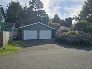 A residential garage with two white doors, surrounded by a gravel driveway, a wooden fence, and lush greenery including a flowering bush. - Cute Rockaway Beach Cabin (Rockaway Beach)