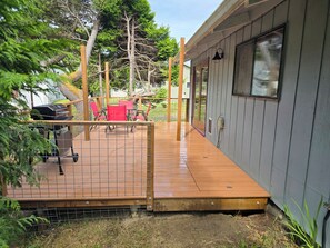 A wooden deck with a grill and chairs, a fenced area, and a building with a window. - Cute Rockaway Beach Cabin (Rockaway Beach)
