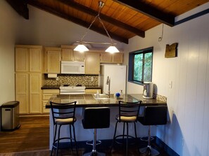 A kitchen with wooden cabinets, a granite countertop, a stainless steel sink, and a bar area with three stools. - Cute Rockaway Beach Cabin (Rockaway Beach)