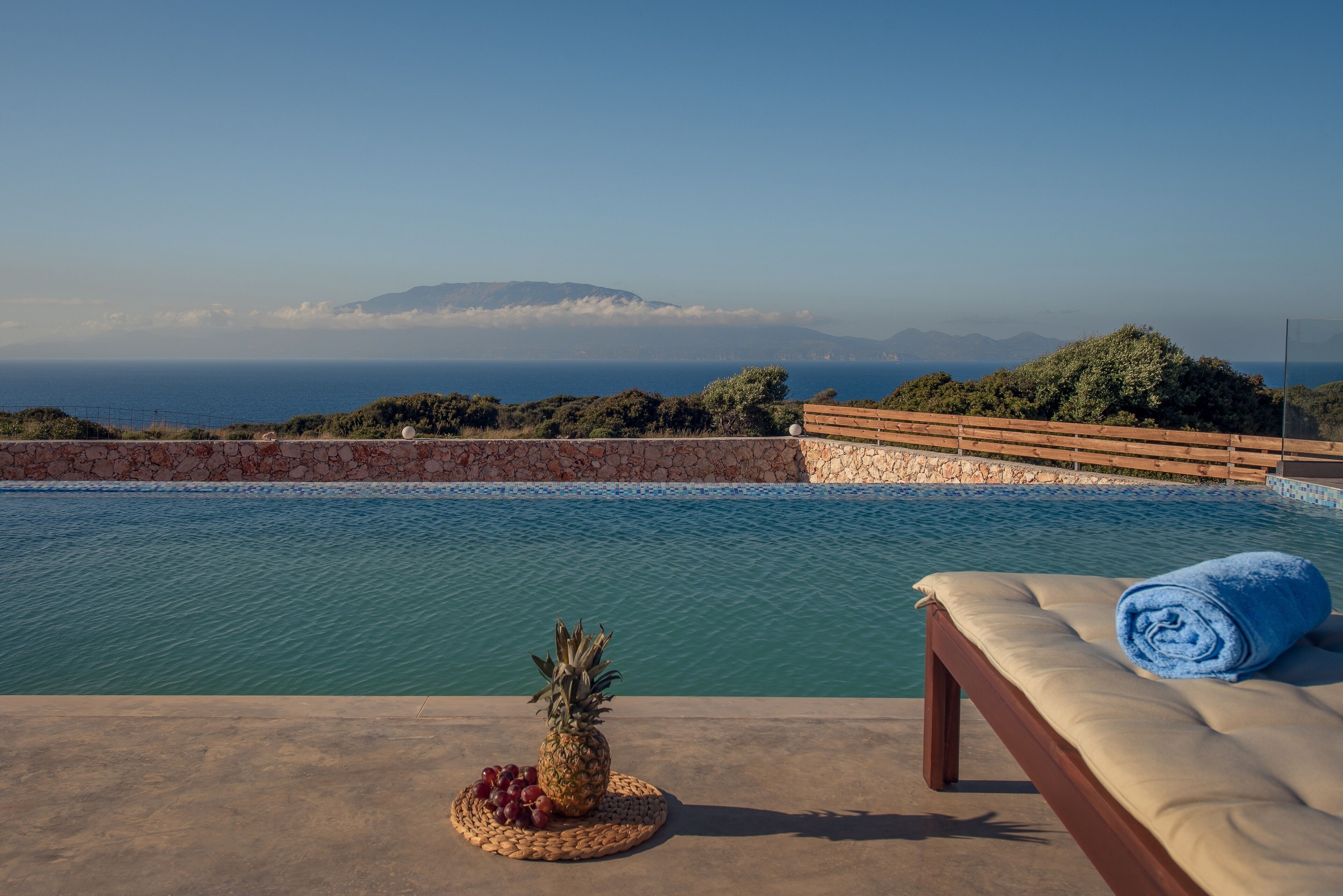 Une piscine bordée d’une terrasse en bois, une chaise longue et, sur le bord, un ananas et des raisins.