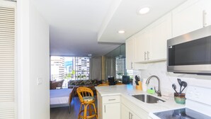 A modern kitchen with white cabinets, a stainless steel microwave, and a built-in oven. There is a dining area with a wicker chair and a small table. - Charming Ilikai Marina Studio, Great Location In Waikiki! (Honolulu)