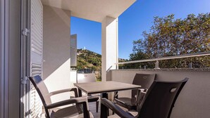 Un balcón al aire libre con una mesa y sillas, barandilla y vistas a una colina con edificios.